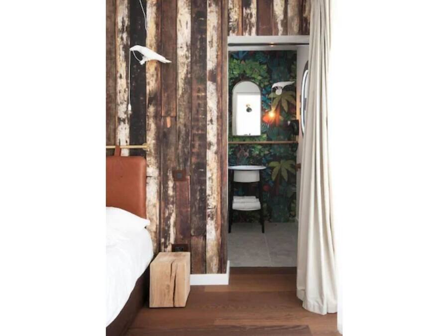 Un dormitorio con cabecera de madera, una mesita de noche pequeña y una mesita auxiliar. Hay una puerta que da a un baño con ducha, lavamanos y toallero.