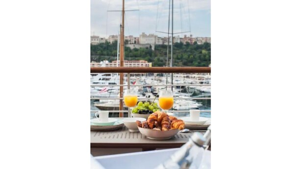 Una mesa de desayuno con cruasanes, un cuenco de fruta y un vaso de zumo de naranja, con vistas a un puerto deportivo con barcos y edificios al fondo.