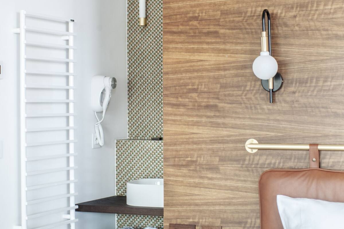 A modern hotel room with a wooden headboard, a white bed, a wooden nightstand with a remote control and glasses, a towel rack, a white hairdryer, and a wall-mounted light fixture.