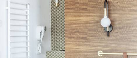 A modern hotel room with a wooden headboard, a white bed, a wooden nightstand with a remote control and glasses, a towel rack, a white hairdryer, and a wall-mounted light fixture.