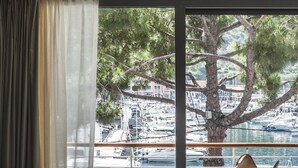Habitación de hotel con cama, una silla, una mesita y vistas a un puerto deportivo a través de amplias ventanas.