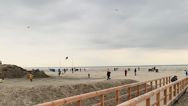 A wooden boardwalk leading to a sandy beach with people walking and flying kites.