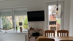 A modern living room with a dining area, large windows, a flat-screen TV, and a fireplace.