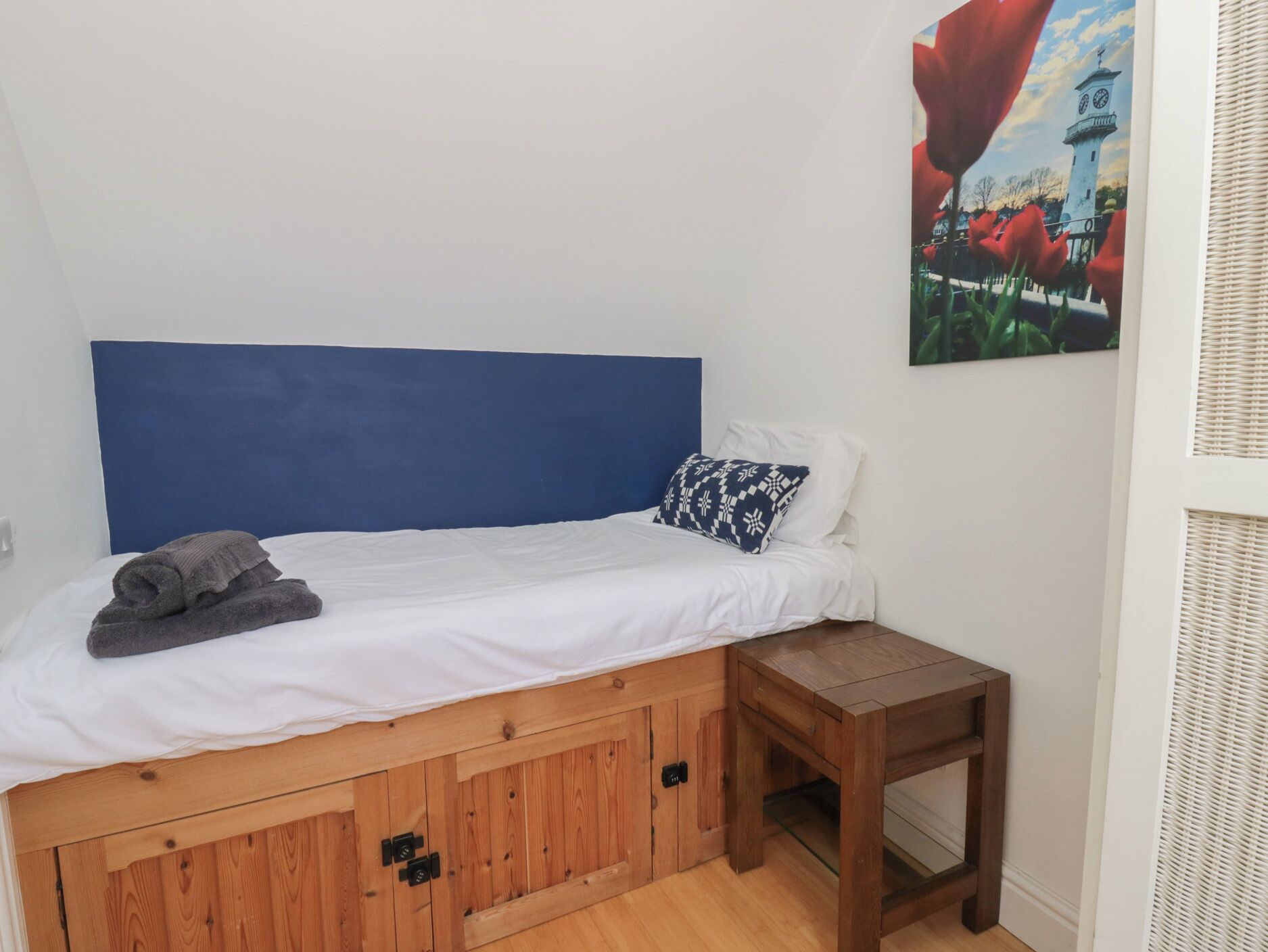 Ein Einzelbett mit blauem Kopfteil, ein kleiner Nachttisch und ein Gemälde eines Leuchtturms.