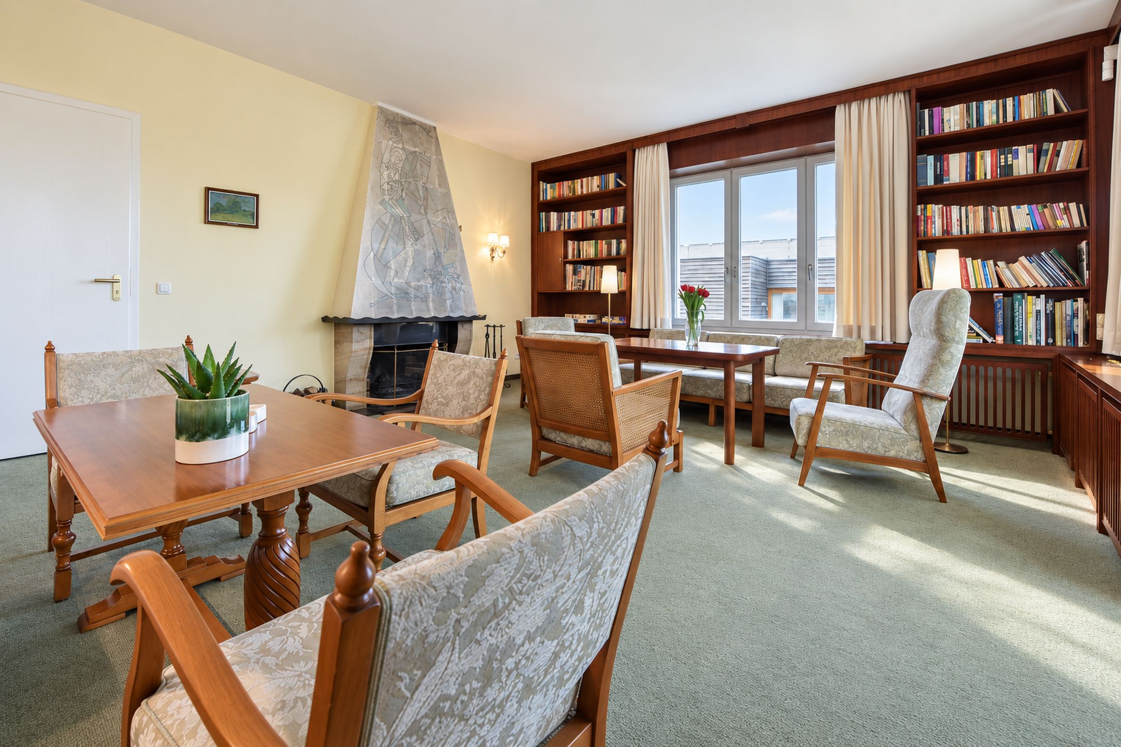 Una habitación con chimenea, una mesa de comedor, sillas, una estantería y una ventana con cortinas.