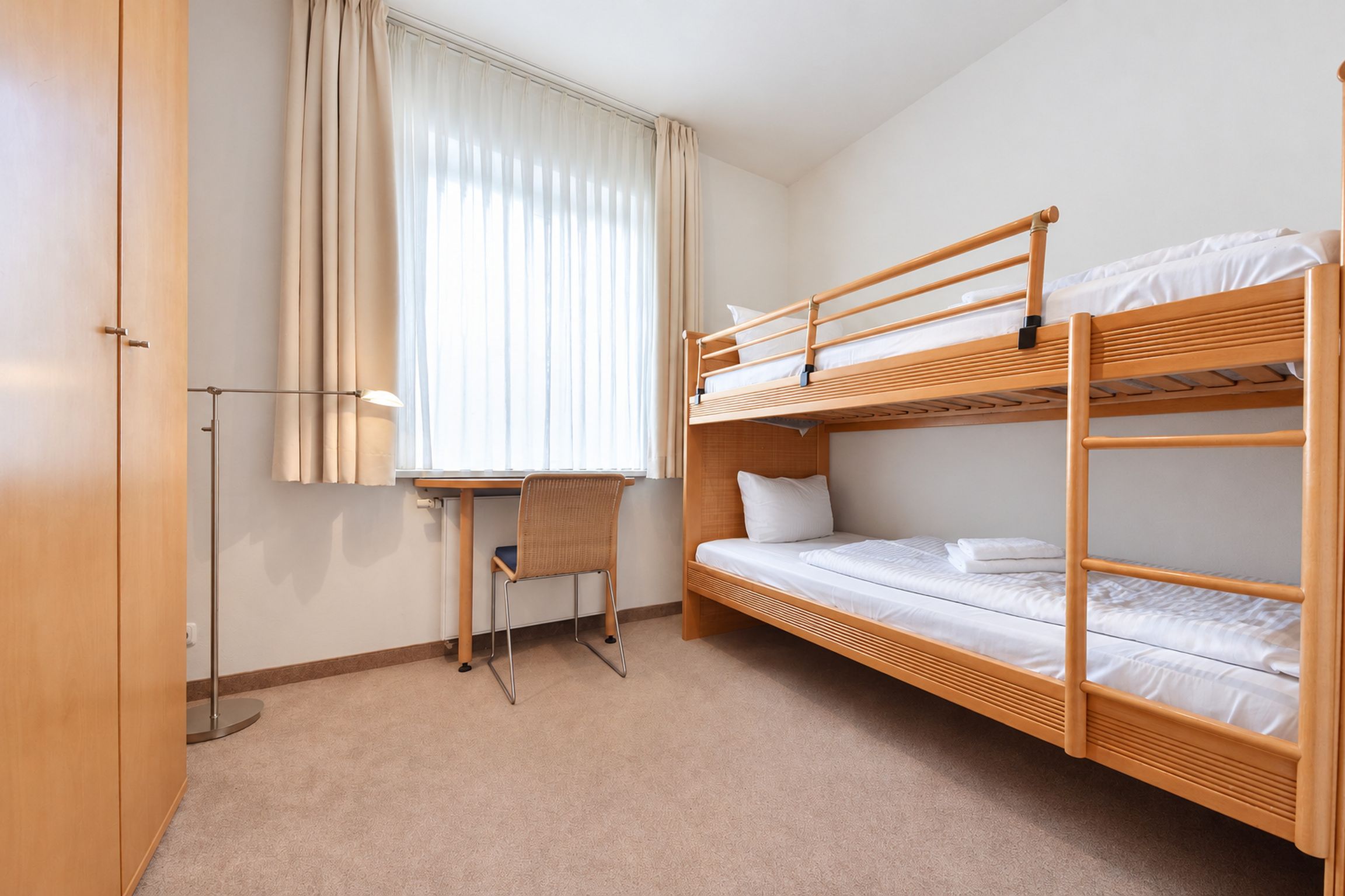A bunk bed room with a desk and chair, a window with curtains, and a wardrobe.