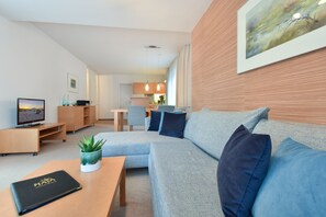 Ein modernes Wohnzimmer mit einem grauen Ecksofa, einem Holztisch und einem Fernseher auf einem Standfuß.