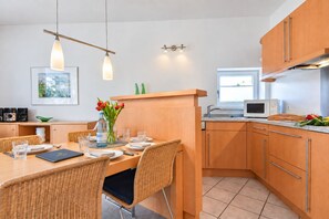 Une cuisine moderne avec des meubles en bois, un coin repas avec des chaises en osier et un petit îlot agrémenté d’un vase de fleurs.
