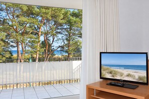 Un balcón con vista a la playa y al océano, un televisor Samsung sobre un atril de madera, y una cortina blanca.