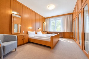 Une chambre d’hôtel avec un grand lit, un coin salon avec un fauteuil, un miroir et une fenêtre avec des rideaux.