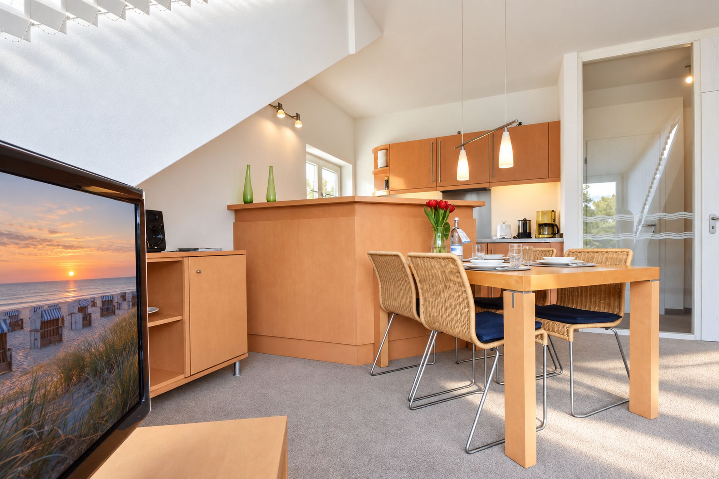 A modern kitchen with wooden cabinets, a dining area with wicker chairs, and a large window overlooking a beach.