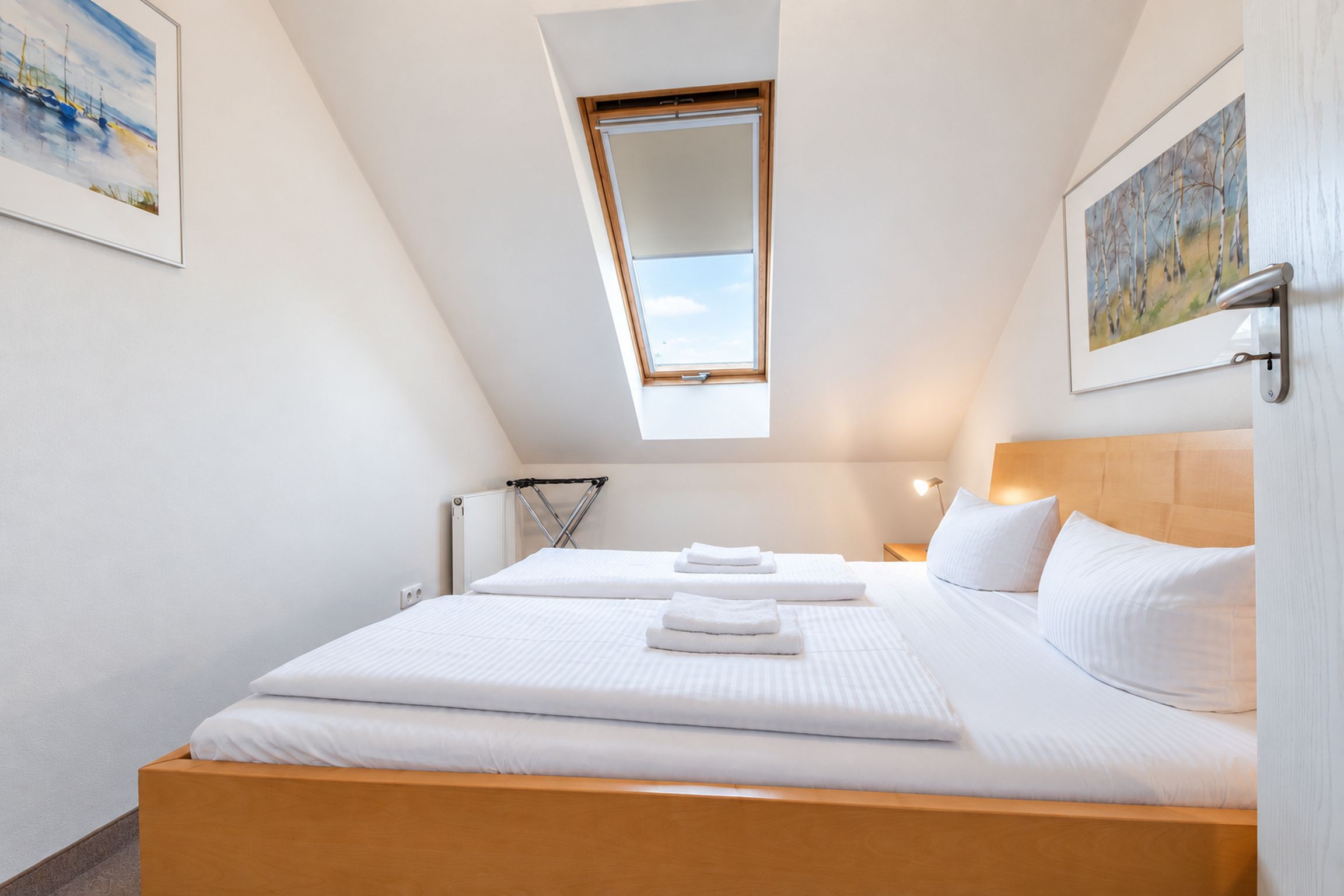 Une chambre avec un plafond en pente, un puits de lumière, un lit avec du linge de maison blanc et un tableau accroché au mur.
