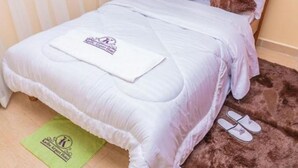 A neatly made bed with a white comforter and a logo on the pillow. There is a pair of slippers on a brown rug beside the bed.
