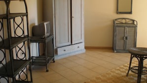 Een kamer met een wit kastje, een televisie op een bijzettafeltje en een stoel met een geweven zitting.