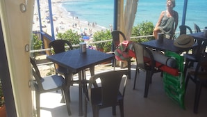 Een balkon met uitzicht op een strand en de zee, met tafels en stoelen, een hoed en een waterfles.