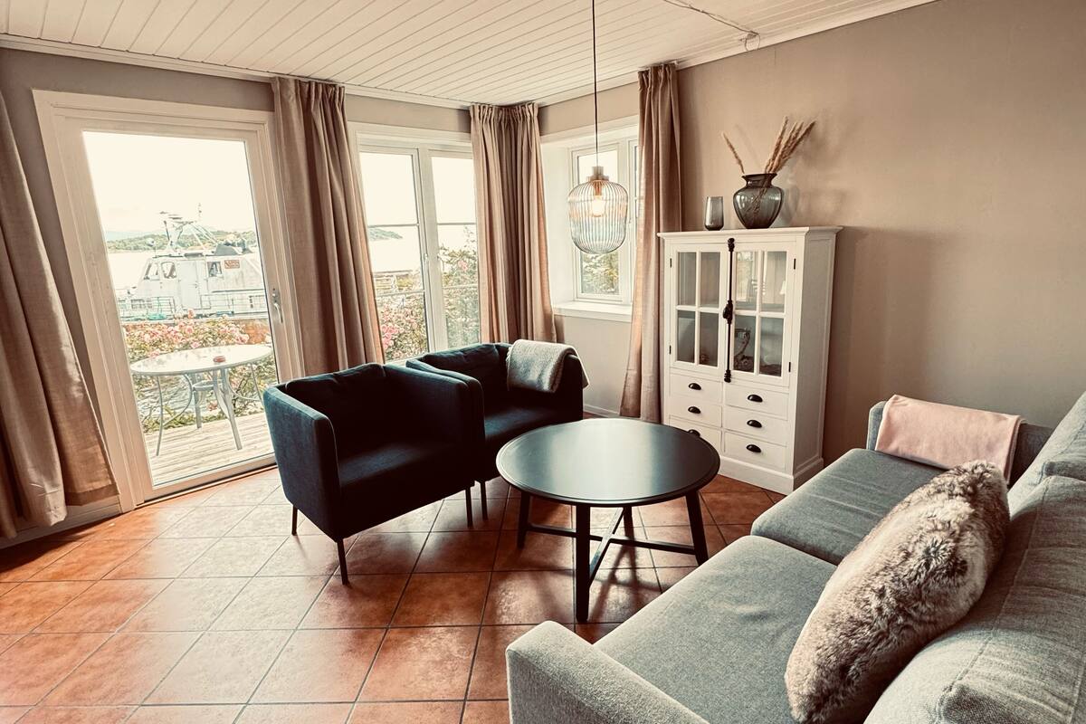 A living room with a sofa, two armchairs, a round table, a cabinet, and a view through a large window.
