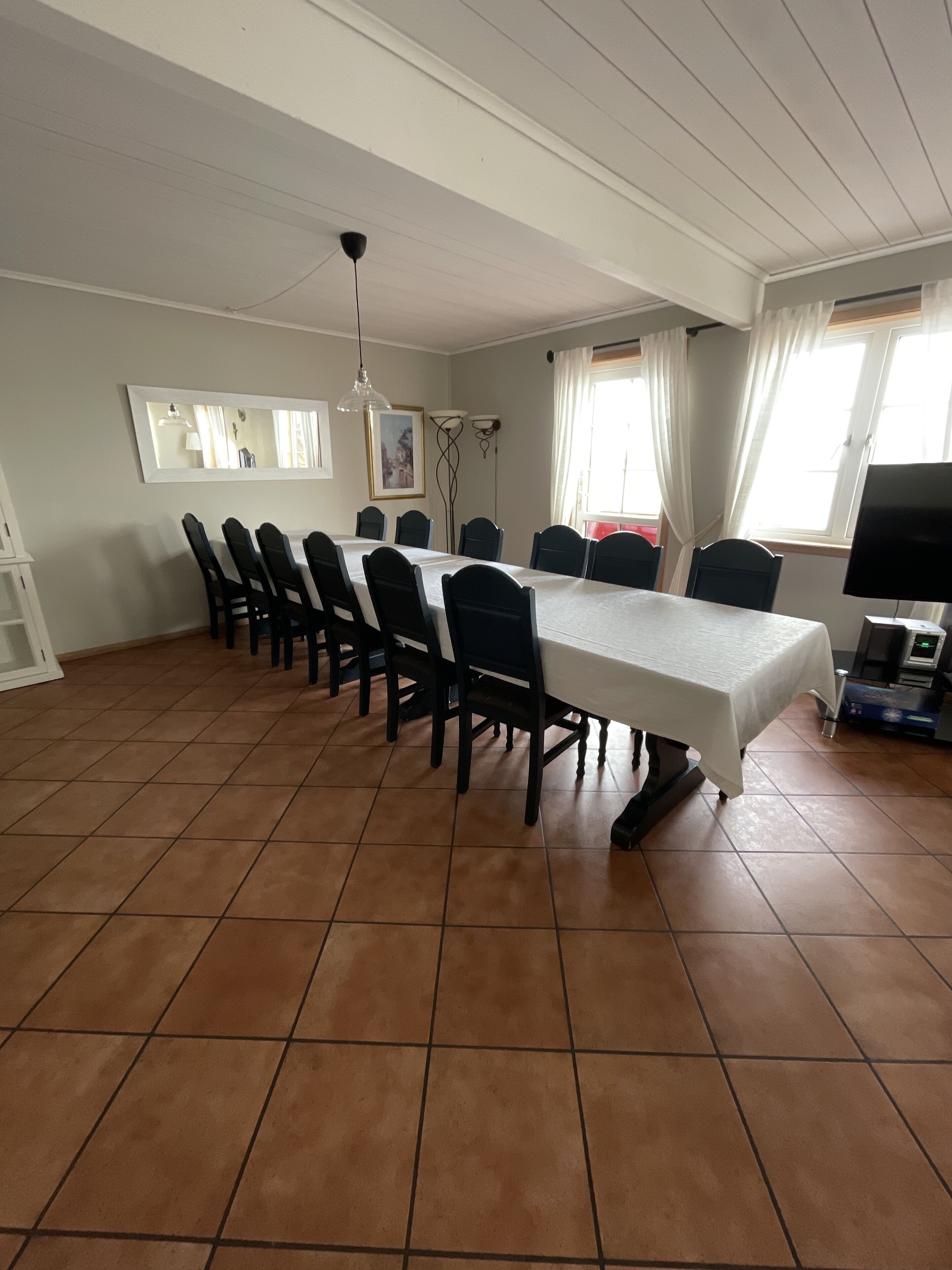 A dining area with a long table, chairs, a TV, and a window with curtains.