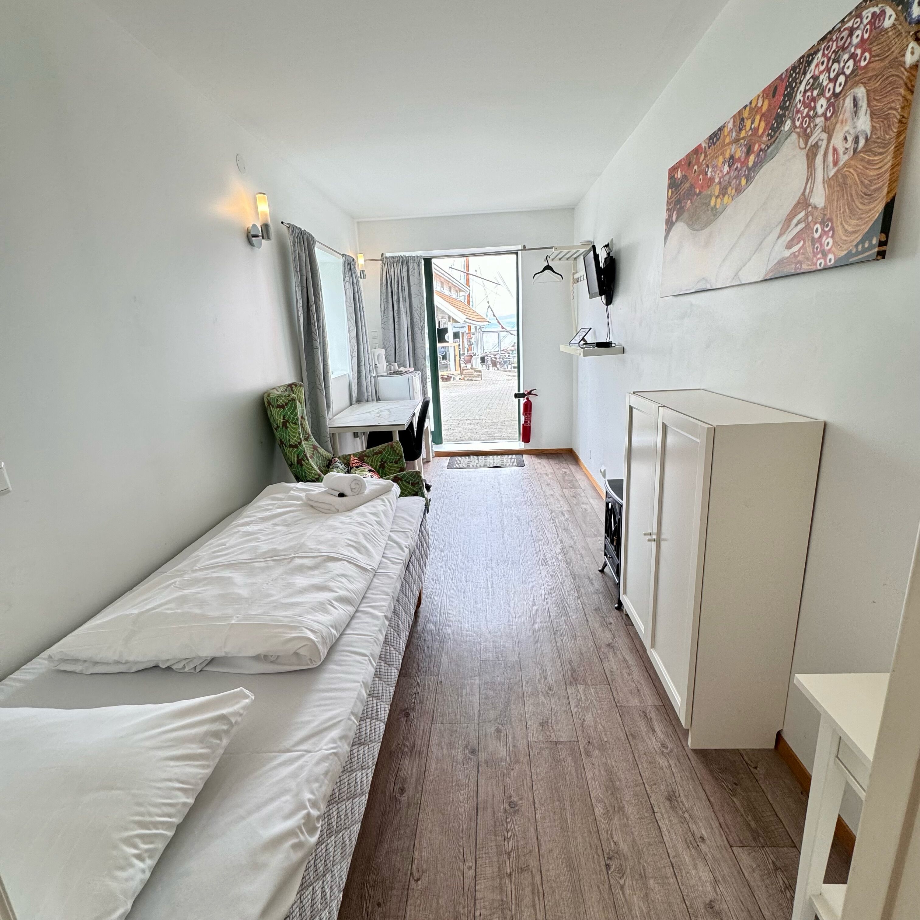 A compact hotel room with a bed, a small desk, a wardrobe, and a painting on the wall.