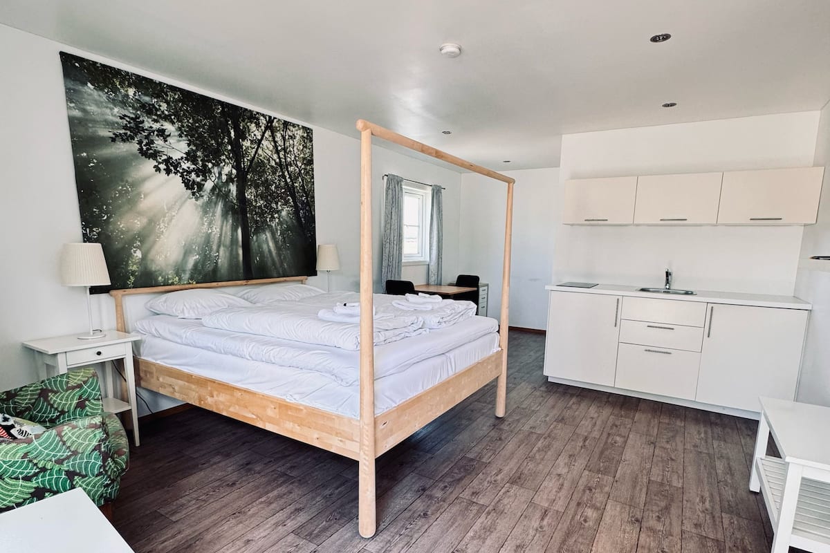 A modern bedroom with a four-poster bed, a large forest-themed tapestry, a desk, and a kitchenette.