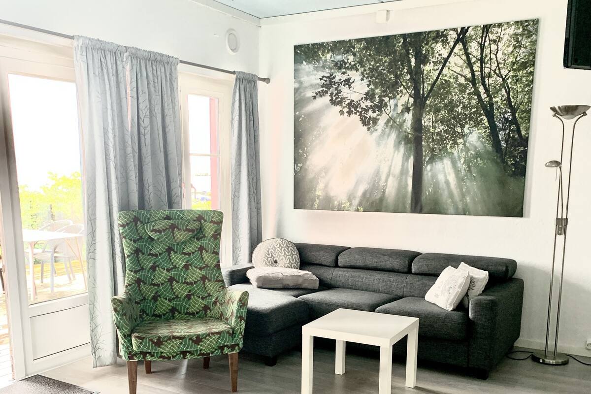 A living room with a green patterned chair, a grey sofa, a white coffee table, and a forest-themed painting on the wall.