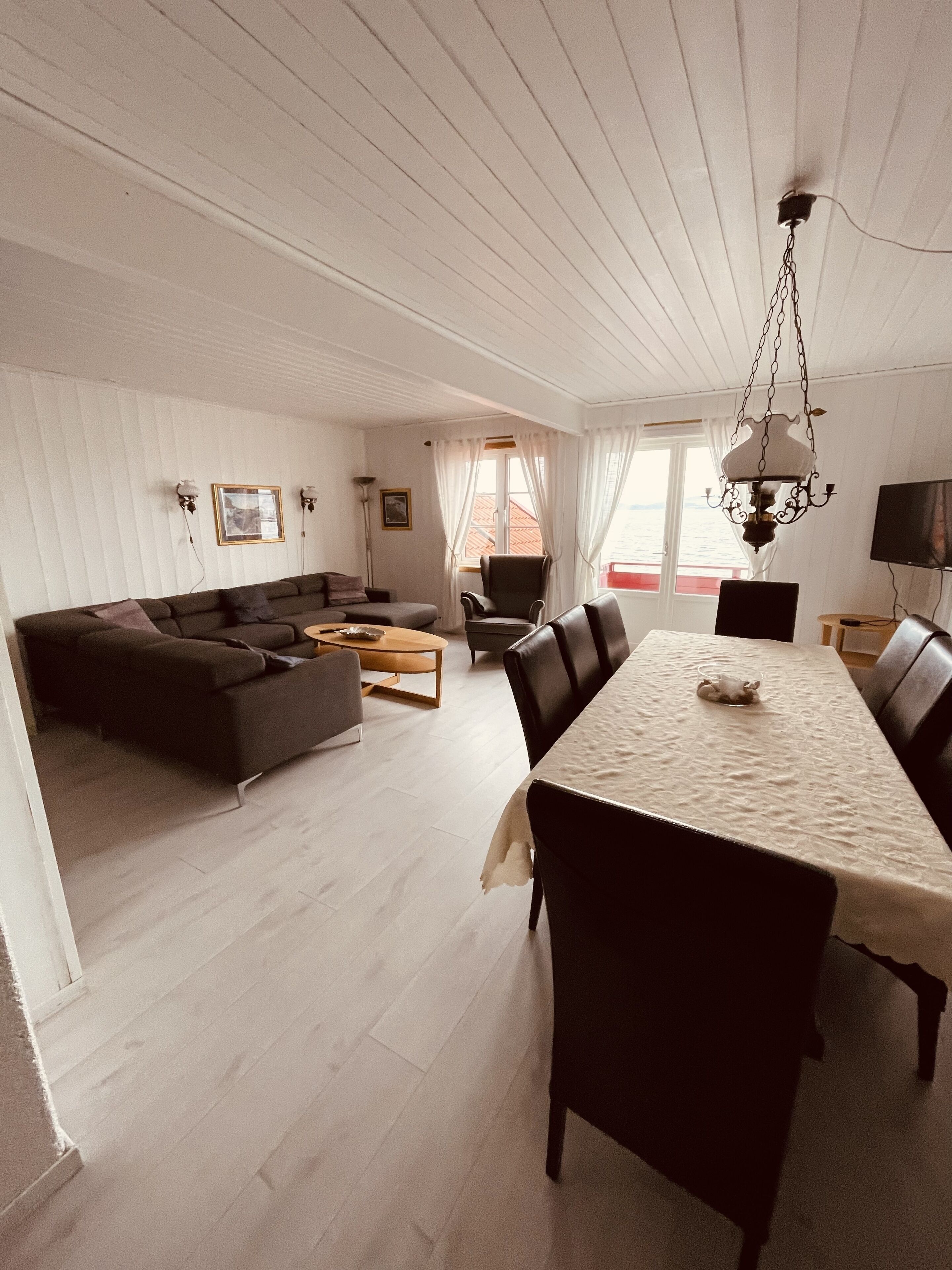 A spacious living area with a sofa, dining table, and chairs. There is a television and a window with curtains.