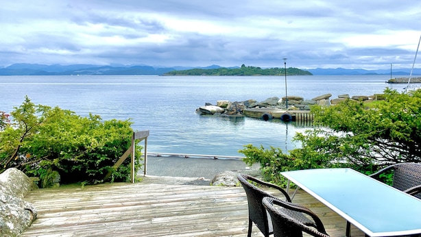 A wooden deck with a table and chairs overlooking a calm body of water, with a rocky breakwater and distant mountains under a cloudy sky.
