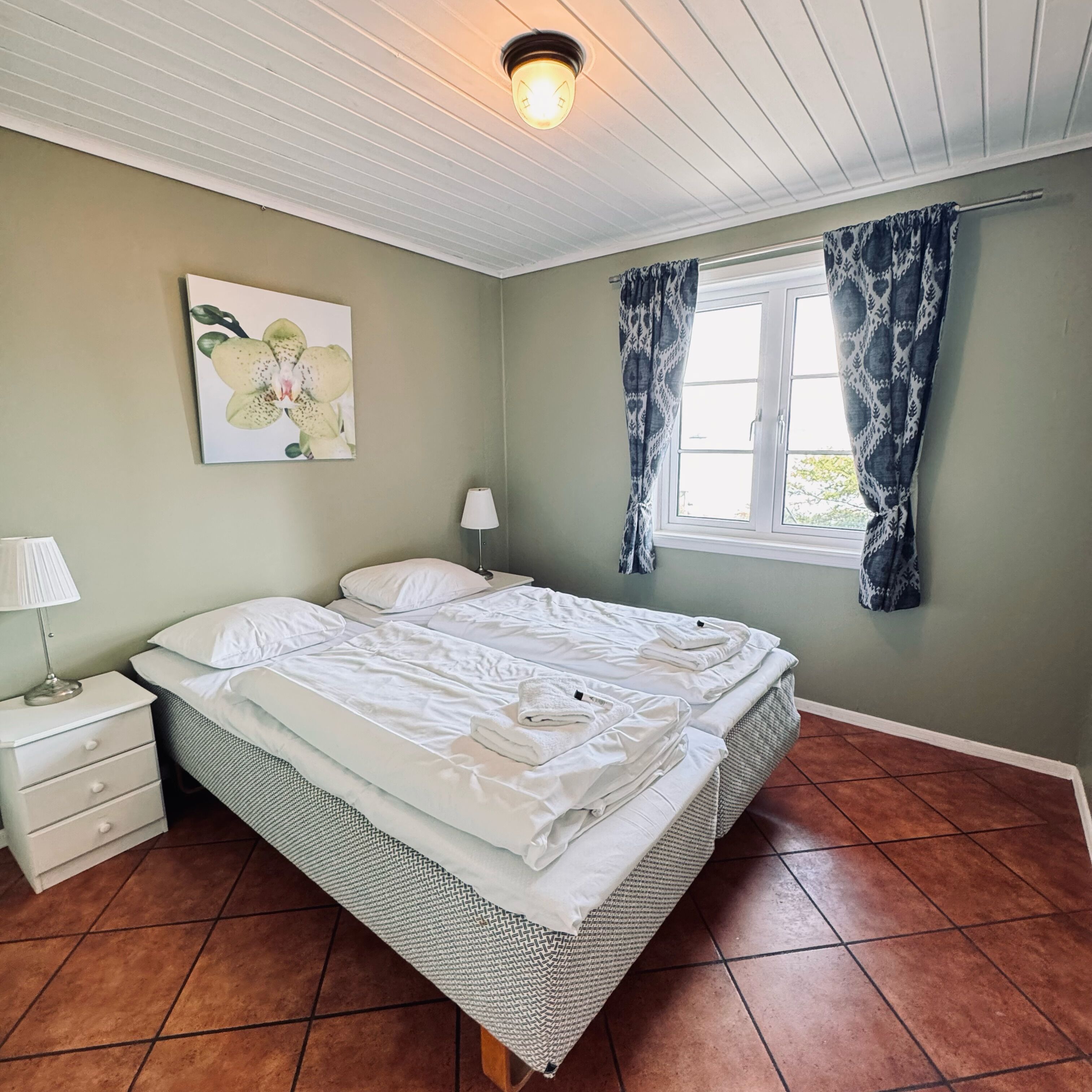 Ein Schlafzimmer mit einem Bett, einem Nachttisch, einer Lampe, einem Fenster mit Vorhängen und einer Deckenleuchte.