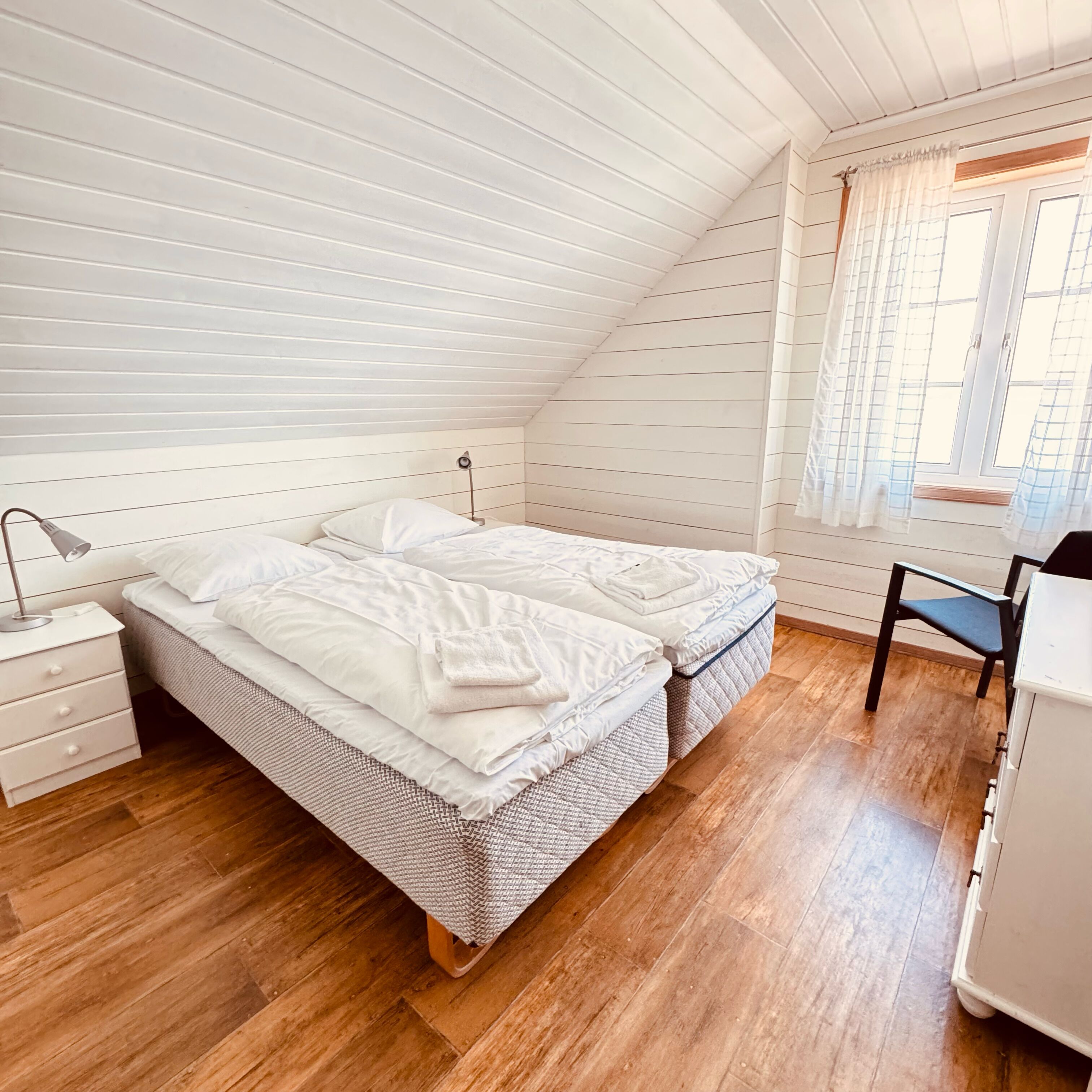 Un dormitorio con techo inclinado, una cama con ropa de cama blanca, una mesita de noche, una silla y una ventana con cortinas.