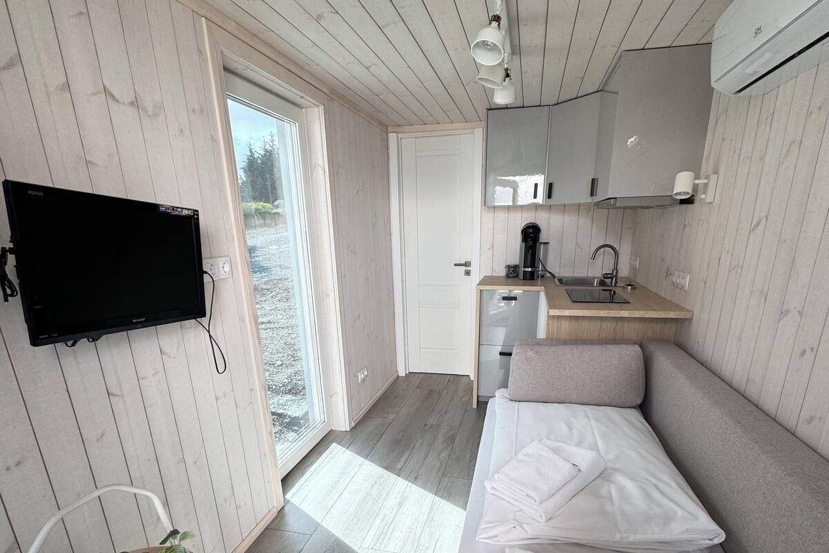A modern room with a bed, a small kitchenette, a television mounted on the wall, and a door leading to an outdoor area.