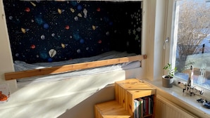 Une chambre d’enfant avec un mur décoré d’un ciel étoilé, un lit en bois, un petit tabouret en bois et une bibliothèque près de la fenêtre.