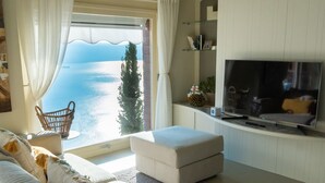 A living room with a sofa, ottoman, and a TV on a stand. There is a window with a view of the sea and a tree. - EnjoyIseoLake - Colosseum - Parzanica (Parzanica)