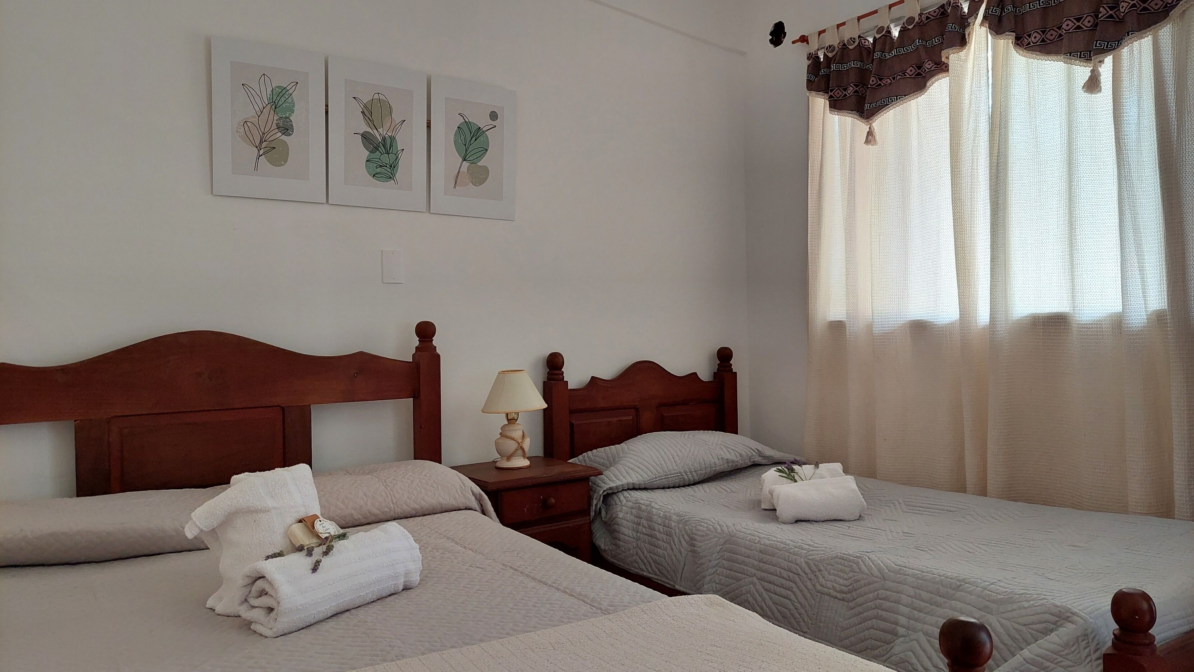 A hotel room with two beds, a nightstand with a lamp, and a window with sheer curtains.