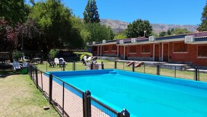 Una piscina con área vallada, un edificio de ladrillo con múltiples ventanas y montañas al fondo.