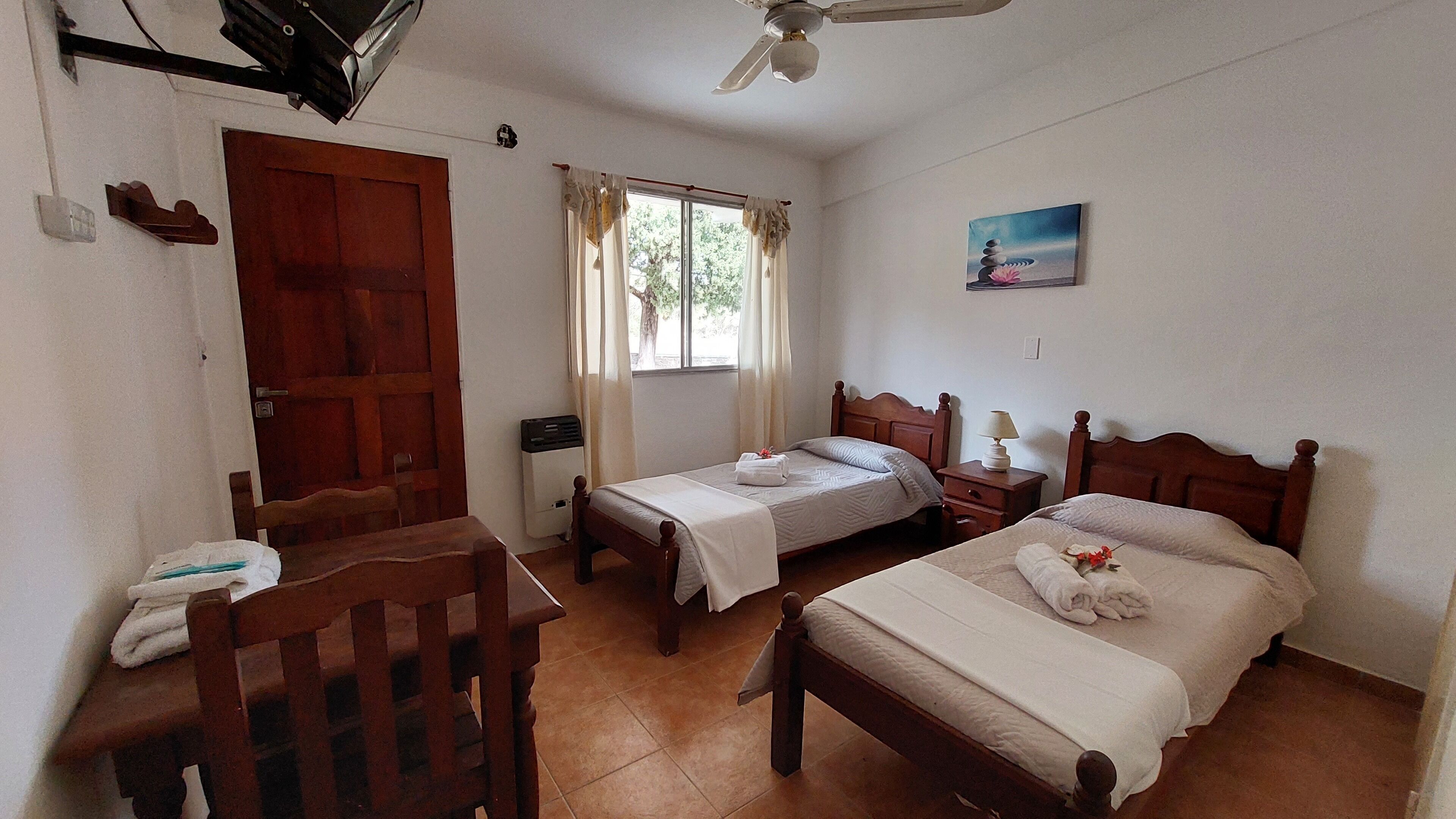 Una habitación con dos camas, una silla de madera, un escritorio, un ventilador de techo, una ventana con cortinas y un cuadro en la pared.