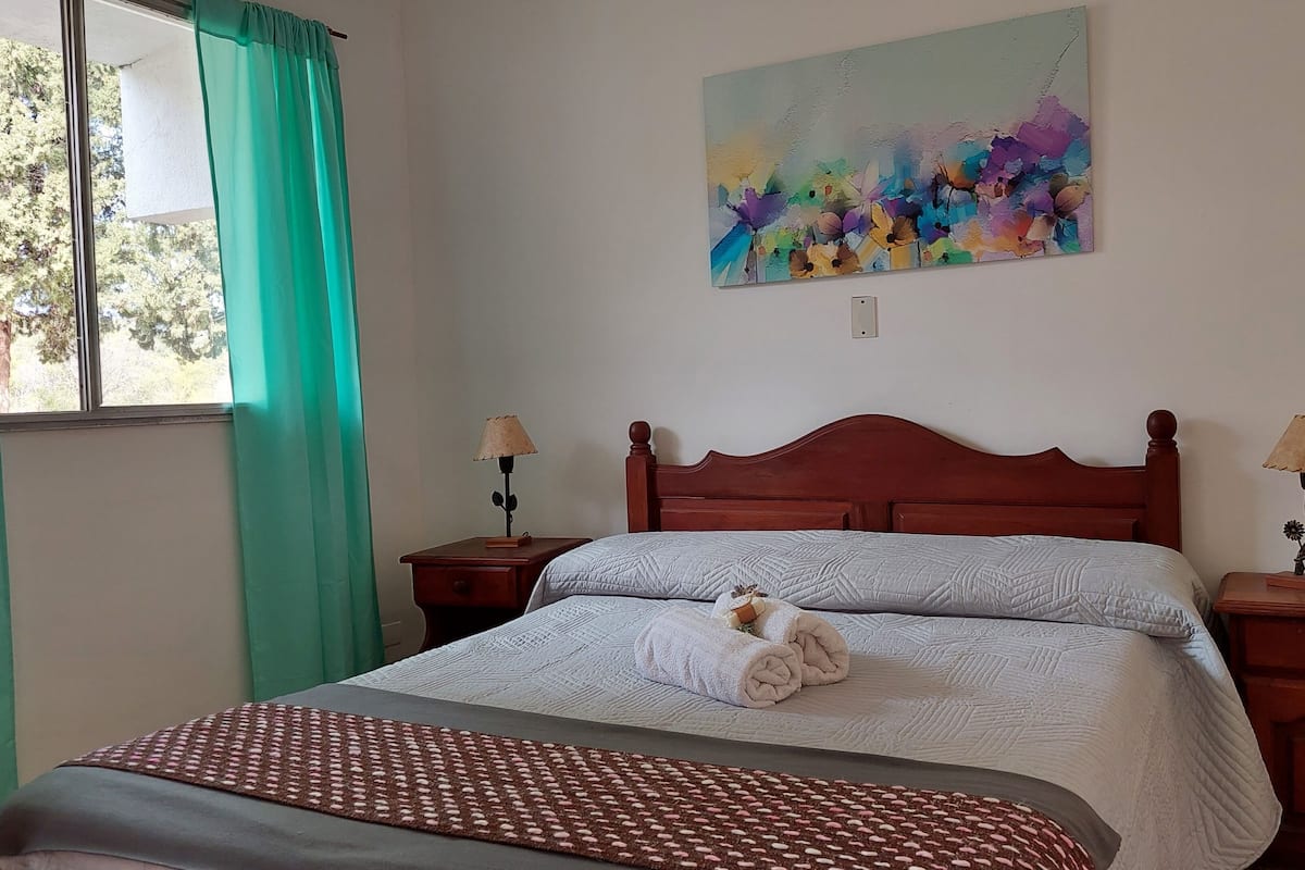 A bedroom with a wooden bed, two bedside tables with lamps, a green curtain, and a colorful abstract painting on the wall.