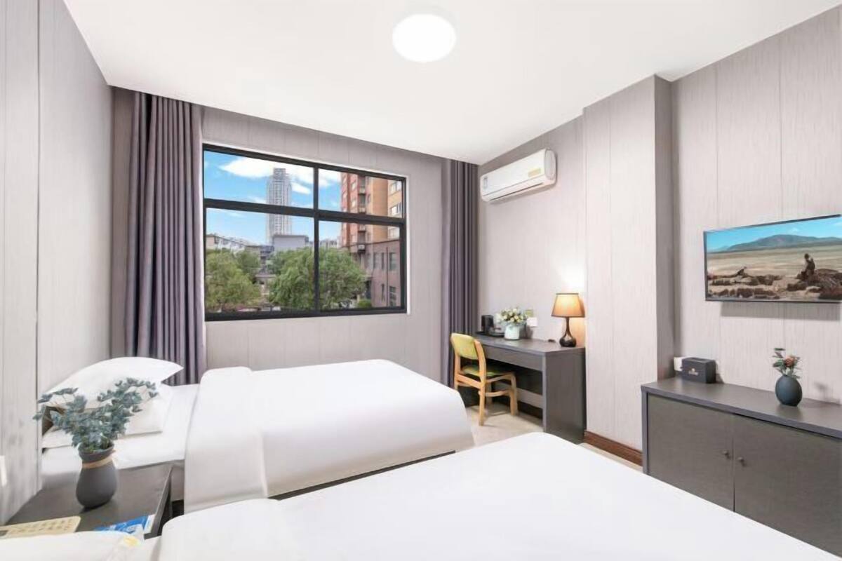 A hotel room with two beds, a desk, a chair, a lamp, an air conditioner, and a painting on the wall.