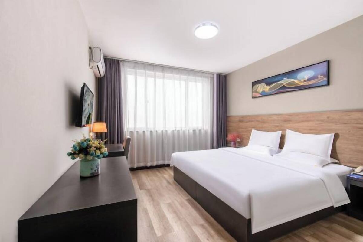 A hotel room with a bed, a desk, a lamp, a vase of flowers, a painting on the wall, and a window with curtains.