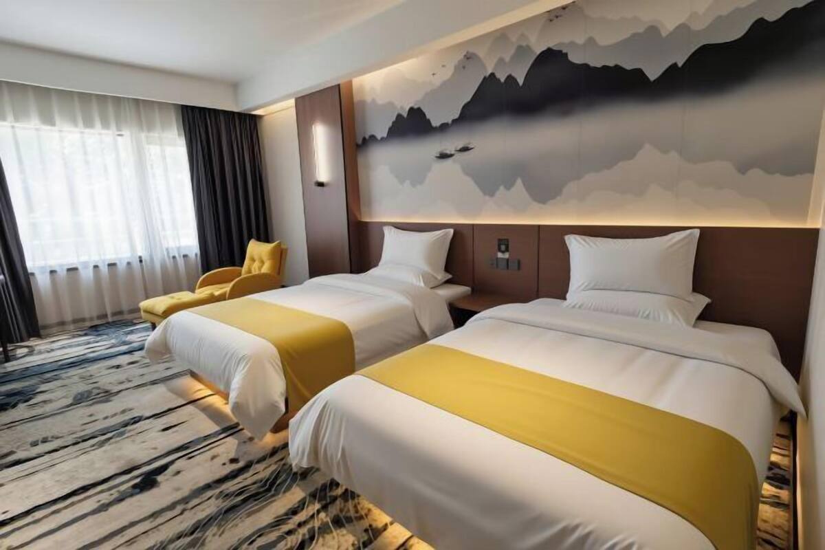 A hotel room with two beds, a large window with curtains, a mountain mural, and a yellow chair.