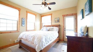 Quarto com cama de madeira, ventilador de teto, janelas amplas e vista para o exterior.