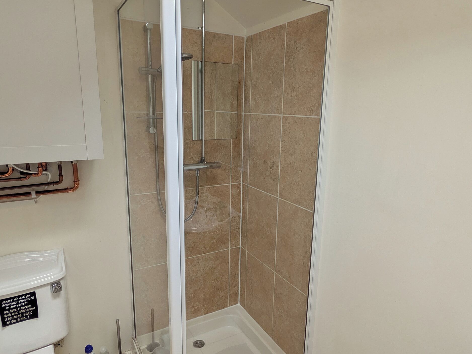 Un baño con cabina de ducha de vidrio, azulejos color beige y tuberías a la vista.