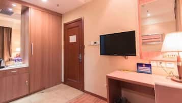 A hotel room with a wooden wardrobe, a television mounted on the wall, a desk with a lamp, and a trash bin.