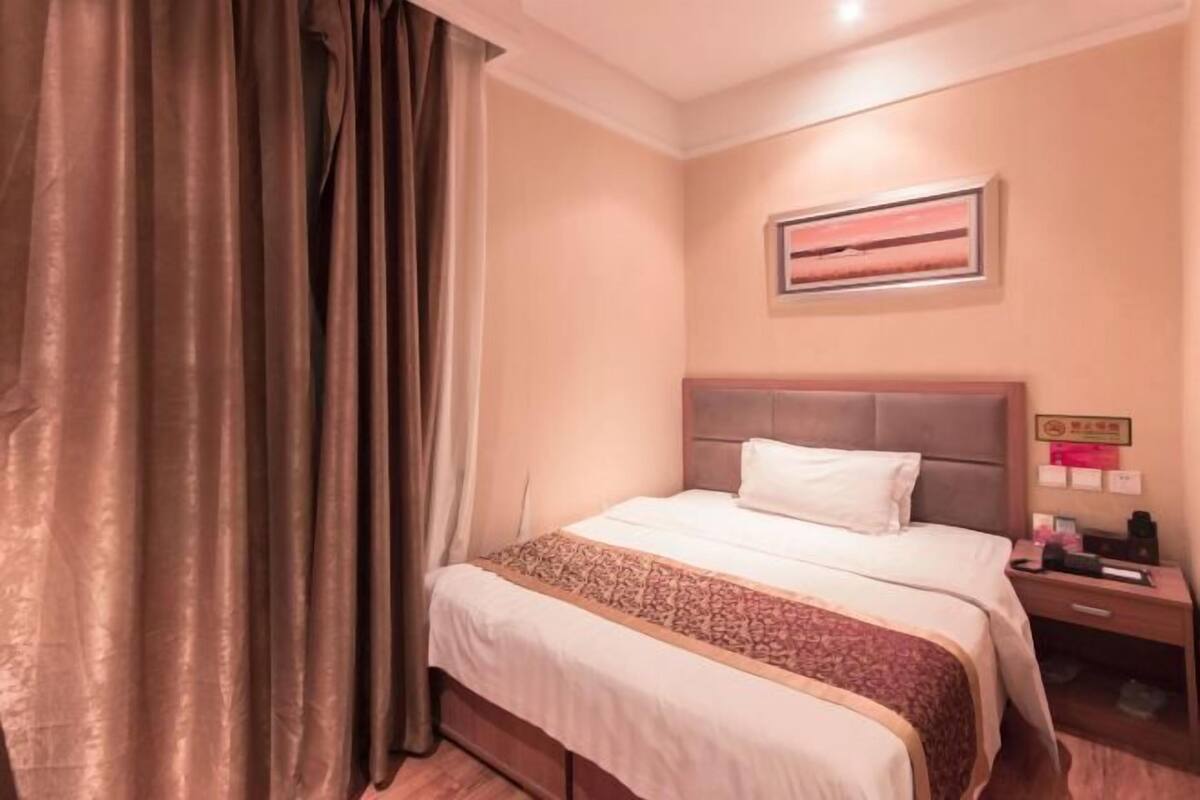 A hotel room with a bed, curtains, a nightstand, a wall-mounted picture, and a small table with items on it.