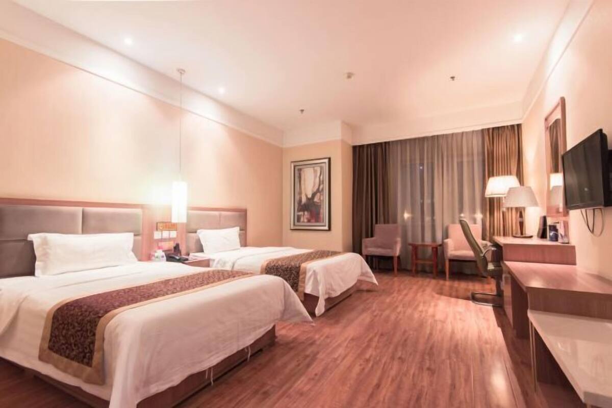 A hotel room with two beds, a desk, a chair, a TV, and a large window with curtains.
