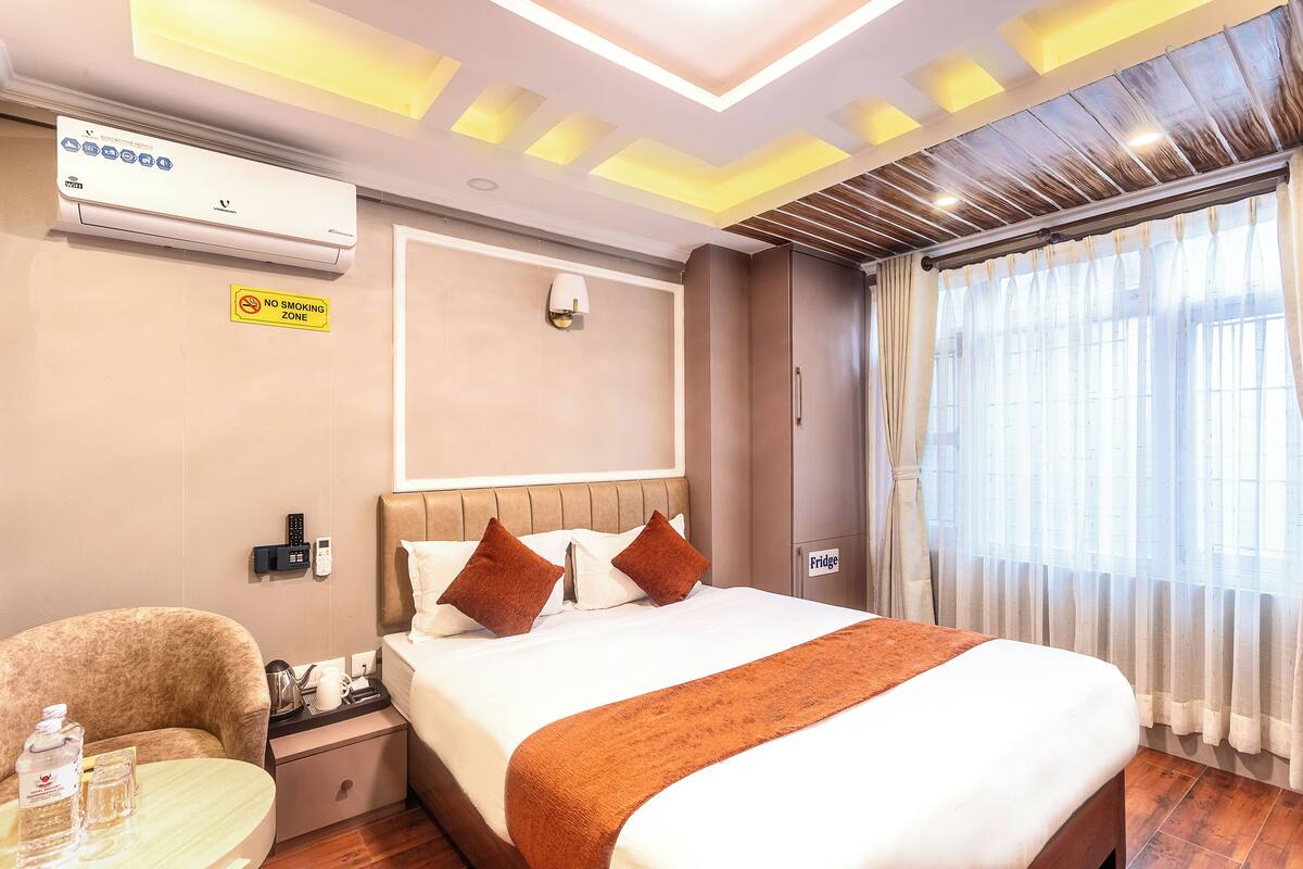 A hotel room with a bed, a bedside table with a phone and a lamp, a chair, a sign indicating a no smoking zone, and an air conditioning unit.