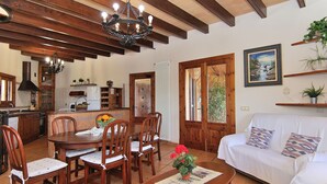 A cozy living room with a white sofa, wooden dining table, and chairs, a chandelier, and a kitchen in the background. - Finca Can Xisco Domatiga 250 Stone villa with swimming pool and high speed internet access (Ariany)