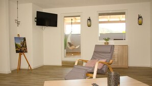 Ein Zimmer mit Holzboden, einem grauen Sessel, einem kleinen Tisch mit Fernbedienung, einem an der Wand befestigten Fernseher und einem Fenster mit Blick.