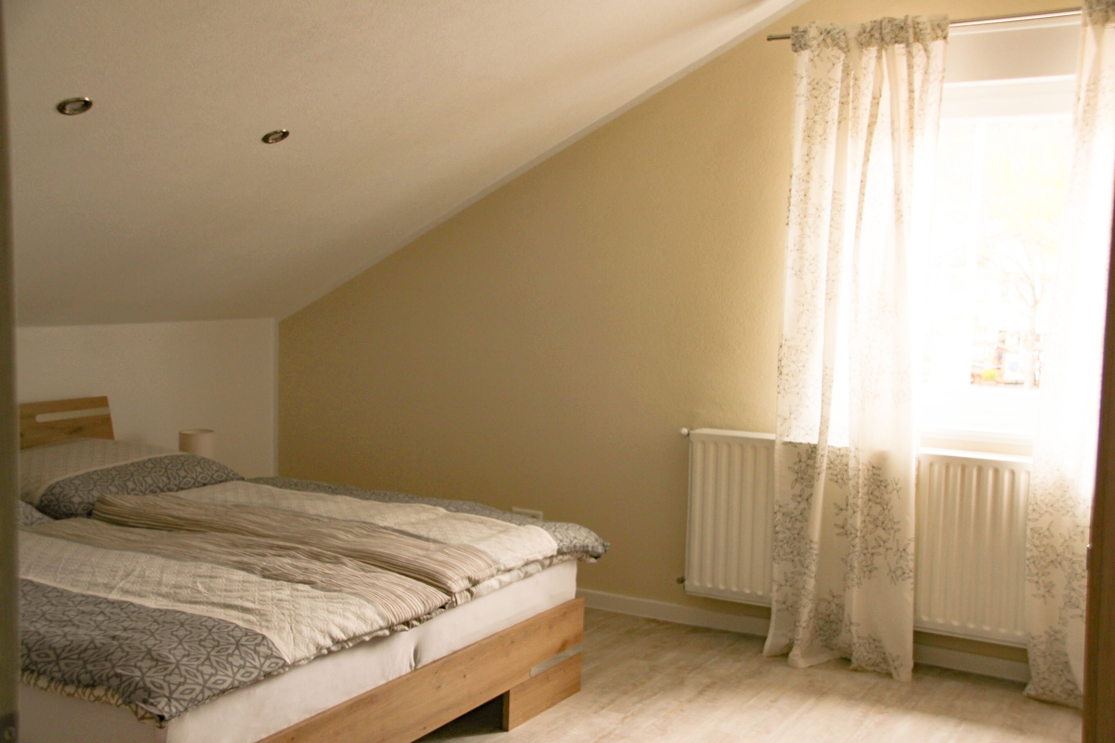 A bedroom with a sloped ceiling, a bed with a wooden frame, a radiator, and a window with sheer curtains.