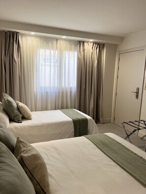 A hotel room with two beds, a window with curtains, a door, and a metal drying rack. - Hotel San Valero Boutique (Zaragoza)
