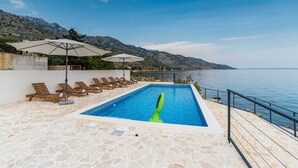 A swimming pool with lounge chairs and umbrellas, surrounded by stone flooring and a view of the sea and mountains. - Luxury Villa Solis (Karlobag)