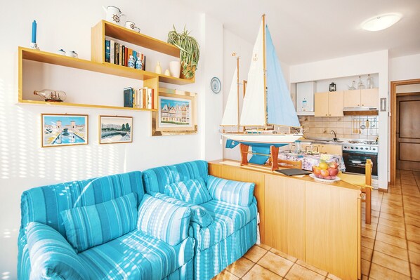 Una sala de estar con un sofá azul a rayas, estanterías de madera con libros y adornos, un modelo de velero, y una cocina con armarios de madera y electrodomésticos.